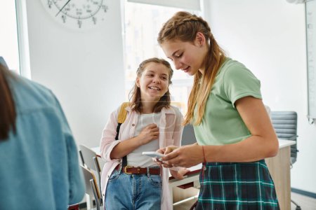 Foto de Compañeros adolescentes positivos usando un smartphone juntos mientras están de pie en el aula en la escuela - Imagen libre de derechos