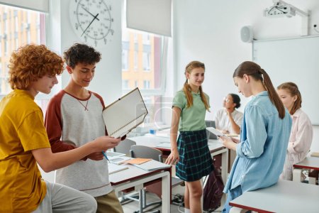 Photo for Positive schoolboys holding notebook near friends and african american teacher in classroom - Royalty Free Image