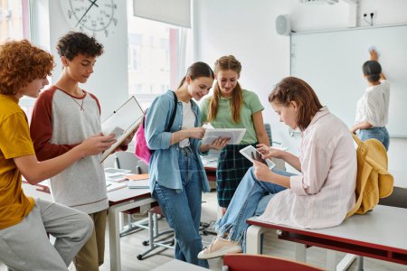 Fröhliche Teenager-Schüler mit Geräten in der Nähe des afrikanisch-amerikanischen Lehrers im Hintergrund im Klassenzimmer