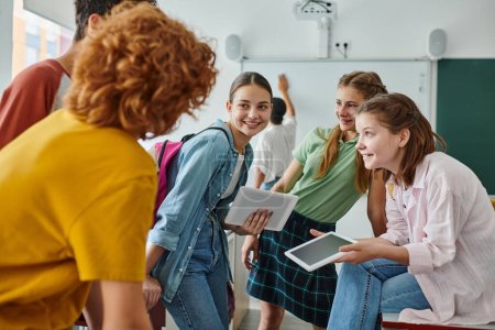 Smiling teen schoolgirls holding digital tablets near blurred friends in classroom in school
