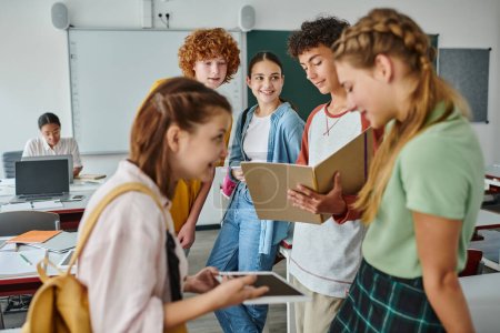 Téléchargez les photos : Un écolier tenant un cahier près de ses amis avec un smartphone et se tenant debout en classe à l'école - en image libre de droit