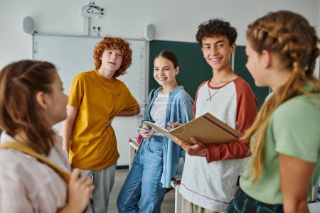 Escolar adolescente sonriente con cuaderno hablando con amigos mientras está de pie en el aula en la escuela