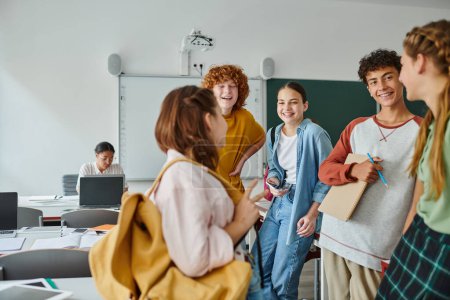 Fröhliche Teenager mit Notizbuch und Smartphone stehen neben verschwommenem Freund im Klassenzimmer