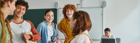 Cheerful teenage classmates with smartphone and notebook standing near friend in classroom, banner