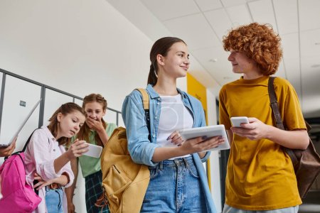heureux adolescent garçon et fille tenant gadgets près de camarades de classe dans le couloir de l'école, concept de retour à l'école