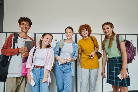 cheerful teenage students holding devices and looking at camera in school hallway, friends