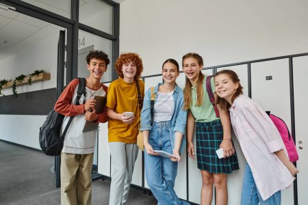 cheerful teenage students holding devices and looking at camera in school hallway, teen friends