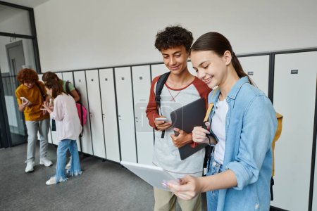 happy boy and girl looking at digital tablet and holding devices in school hallway, teen classmates