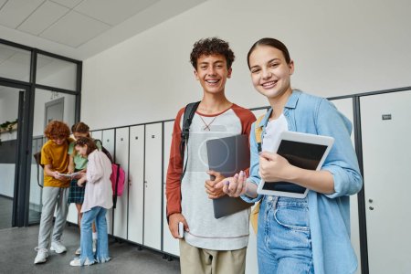Foto de Feliz adolescente compañeros de clase sosteniendo dispositivos y mirando a la cámara en el pasillo de la escuela, estudiantes adolescentes - Imagen libre de derechos
