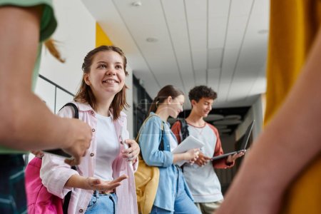 Foto de De vuelta a la escuela, chica adolescente feliz hablando con compañeros de clase en el pasillo, estudiantes borrosos utilizando dispositivos - Imagen libre de derechos