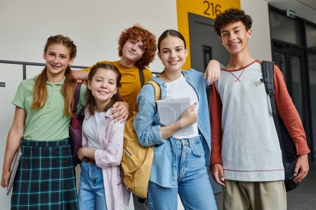 Foto de De vuelta a la escuela, adolescentes felices mirando la cámara en el pasillo, sosteniendo los dispositivos y sonriendo juntos - Imagen libre de derechos