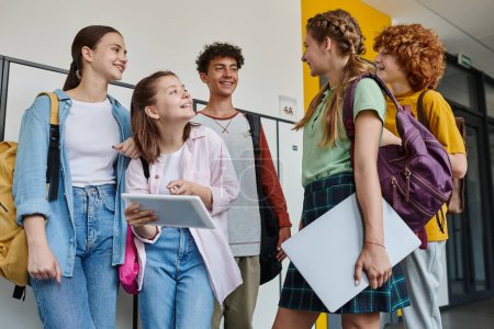 Foto de Adolescente apuntando a la tableta digital y mirando a compañeros de clase, estudiantes adolescentes en el pasillo de la escuela - Imagen libre de derechos