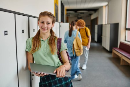 positive teen girl holding laptop and looking at camera in hallway, back to school concept