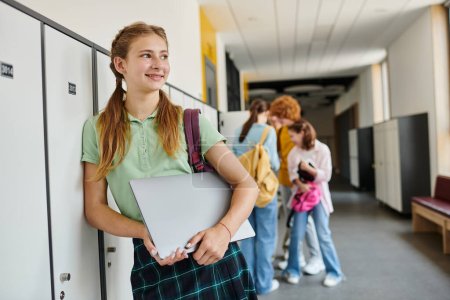 Foto de Chica adolescente positiva sosteniendo el ordenador portátil y mirando hacia otro lado en el pasillo, de vuelta al concepto de la escuela, edad moderna - Imagen libre de derechos