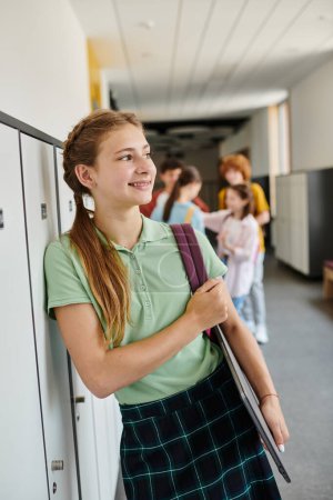 cheerful teenage girl holding laptop and looking away in hallway, back to school concept, modern age