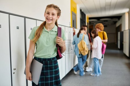fröhliches Teenager-Mädchen hält Laptop in der Hand und schaut im Flur weg, verschwommen, Schulkinder im Hintergrund