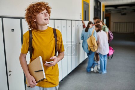 alegre pelirroja colegial usando smartphone y sosteniendo portátil, desenfoque, estudiantes en el pasillo de la escuela