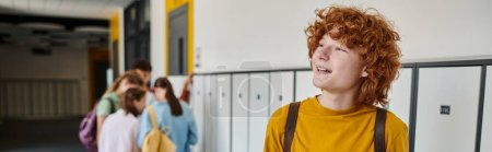 banner, redhead and curly schoolboy looking away while smiling in school hallway, blurred, students