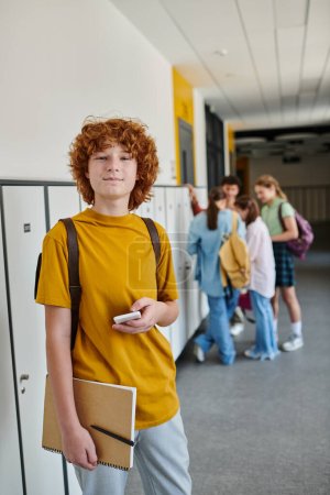 redhead schoolboy holding smartphone, happy student in school hallway looking at camera during break