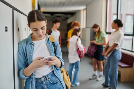 Téléchargez les photos : Adolescente textos sur smartphone dans le couloir de l'école, les étudiants et l'enseignant sur fond flou - en image libre de droit