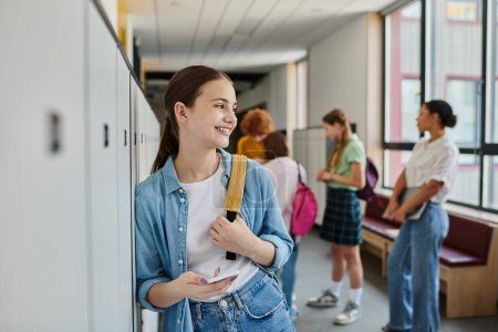 happy teenage girl holding smartphone in school hallway, cultural diversity, teacher and kids, blur
