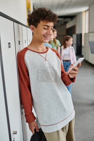 Téléchargez les photos : Heureux écolier avec appareil dentaire en utilisant smartphone dans le couloir de l'école, étudiant pendant la pause, les médias sociaux - en image libre de droit