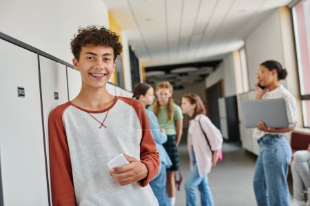 happy boy with braces holding smartphone and looking at camera during break in school hallway