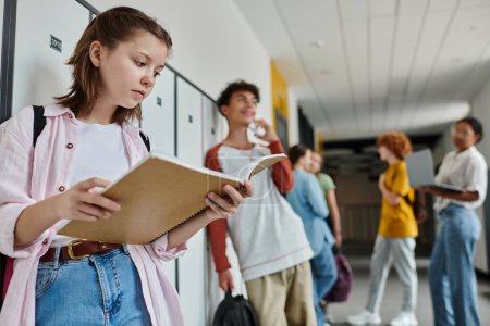 Foto de Colegiala sosteniendo cuaderno y de pie cerca borrosa y feliz compañero de clase adolescente, estudiantes, pasillo - Imagen libre de derechos