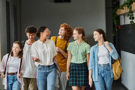 Photo for Cheerful african american teacher walking with teenage students in hallway of school, education - Royalty Free Image