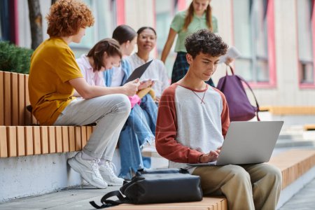 back to school, smart teenage boy using laptop near classmates outdoors, diversity, students