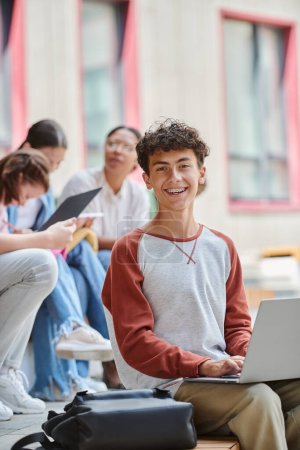 back to school, happy teenage boy with braces using laptop near blurred teacher and girls, diversity