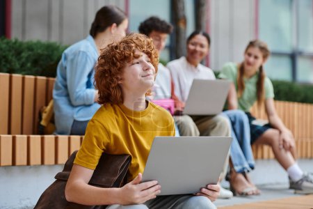 garçon rousse rêveuse avec les cheveux roux bouclés souriant et tenant ordinateur portable, pause, flou, diversité, étudiants