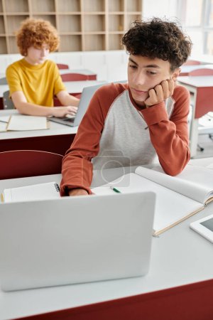 ennuyé garçon bouclé à l'aide d'un ordinateur portable en classe, concept de retour à l'école, e-learning, affectation, camarades de classe