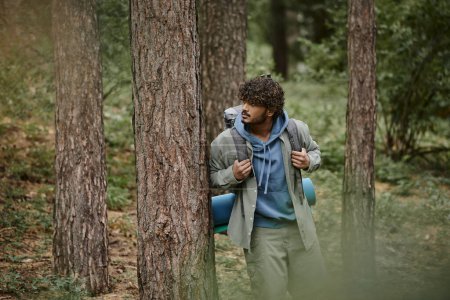 lockiger junger indischer Reisender mit Rucksack, der in der Nähe eines Baumes im Wald steht
