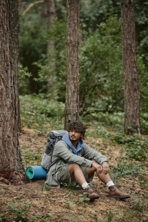 lockiger junger indischer Wanderer mit Rucksack und Trekkingstöcken, der sich in der Nähe von Bäumen im Wald ausruht