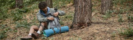 jeune voyageur indien bouclé ouvrant sac à dos tout en se reposant et assis sur le sol dans la forêt, bannière