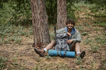 joyeux jeune homme indien regardant la caméra près du sac à dos sur le sol dans la forêt