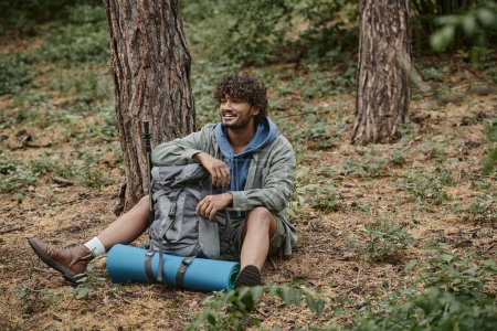 despreocupado joven turista indio descansando mientras está sentado cerca de la mochila en el suelo en el bosque