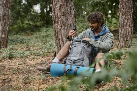 Téléchargez les photos : Jeune randonneur indien positif prenant des bâtons de trekking du sac à dos sur le sol dans la forêt - en image libre de droit