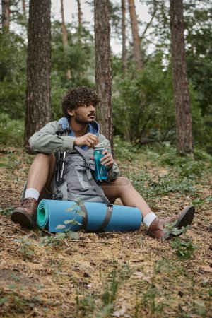 young indian traveler holding sports bottle near backpack with trekking poles in forest