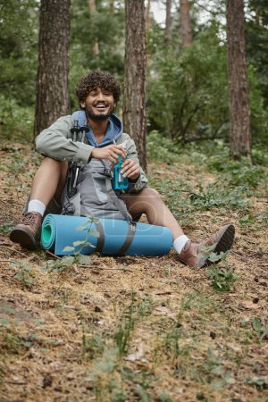 senderista indio despreocupado sosteniendo botella deportiva cerca de la mochila y bastones de trekking en el suelo en el bosque