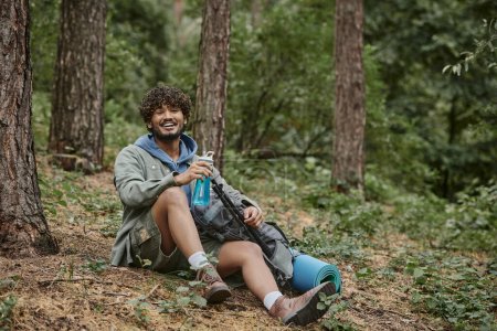 joyeux jeune touriste indien tenant bouteille de sport près du sac à dos sur le terrain dans la forêt