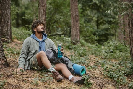 relaxed young indian tourist holding sports bottle near backpack while sitting on ground in forest