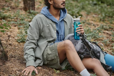 cropped view of young indian man holding sports bottle while sitting near backpack in forest