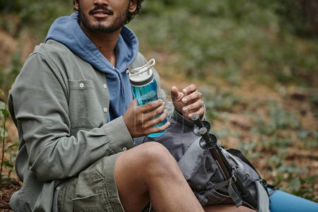 cropped view of indian traveler holding sports bottle near backpack in blurred forest