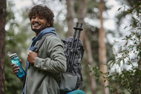 cheerful indian tourist with backpack and sports bottle looking at camera in blurred forest