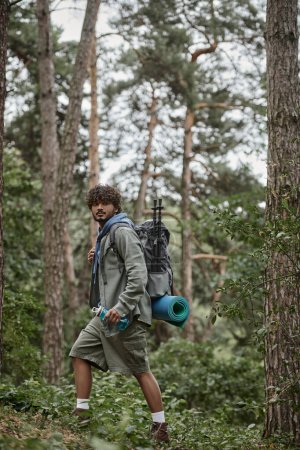 curly indian traveler holding backpack and sports bottle while walking in forest