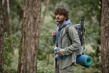 curly young indian tourist in shirt holding backpack and sports bottle in forest