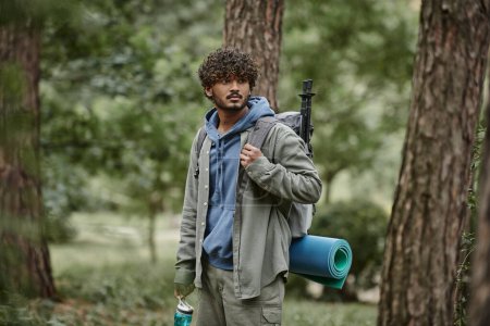 young indian traveler in shirt holding backpack and sports bottle near tees in forest