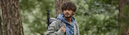 Téléchargez les photos : Jeune touriste indien bouclé avec sac à dos et bâtons de trekking regardant loin dans la forêt, bannière - en image libre de droit